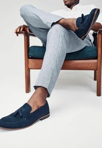 Mocasines de ante azul marino con detalles de borlas, tacón plano y suela marrón contrastante, llevados con pantalones de cuadros gris claro.
