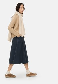 Donkerblauwe spijkerrok tot op de knie gecombineerd met een beige gebreide trui en een crèmekleurige sjaal; bruine sneakers met witte zolen; ontspannen pasvorm en casual stijl.