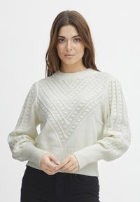 Pull en tricot couleur crème avec un design texturé et des motifs géométriques, des poignets côtelés et un col rond. Tissu doux avec une forme structurée.