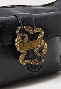 Sac en cuir noir avec un fermoir en métal doré en forme de serpent, orné d'une plaque logo en relief indiquant "JUST CAVALLI MILANO." Texture lisse.