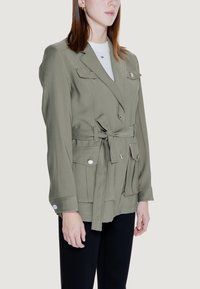 Trench coat verde oliva con design doppiopetto, vita cinta, numerose tasche e sottili dettagli in bottone. Tessuto morbido e vestibilità affusolata.
