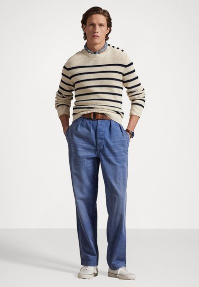 Cremfarbener und navygestreifter Strickpullover mit rundem Hals, kombiniert mit hellblauen, plissierten Hosen und beigen Bootsschuhen.