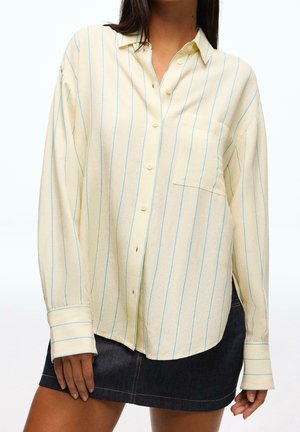 Femme portant une chemise ample crème à boutons avec de fines rayures verticales bleues et une mini-jupe en denim foncé.