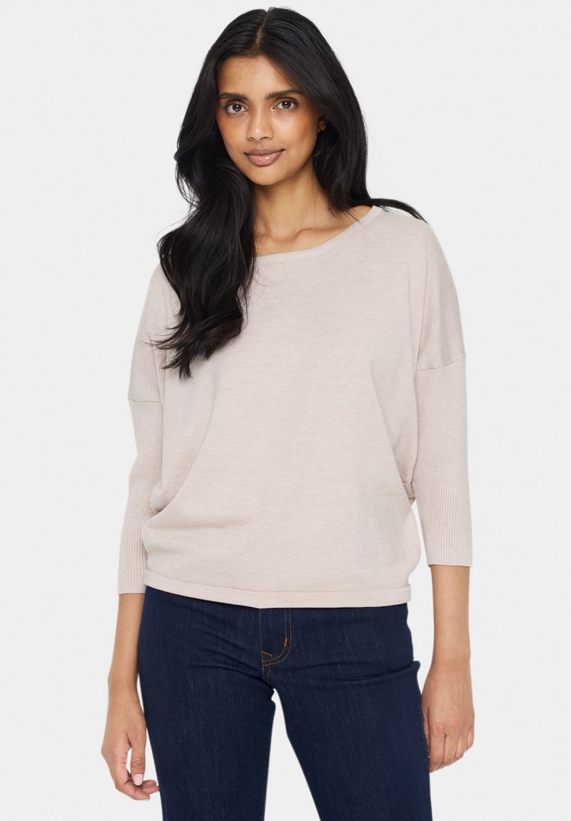 Hellbeige Pullover mit entspannter Passform, Boot-Ausschnitt und Dreiviertelärmeln. Rippstruktur an den Ärmeln und dem Saum. Getragen mit dunklen Jeans.