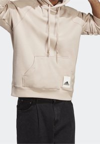 Beige huppari, jossa on etutasku, ribbireunat hihansuissa ja helmassa. Alhaalla vasemmassa kulmassa Adidas-logo. Pehmeä rakenne.