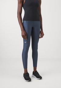 Navy leggings yang terbuat dari kain elastis, desain pas, menampilkan logo putih kecil. Dipasangkan dengan sepatu atletik hitam dengan tali bertekstur.