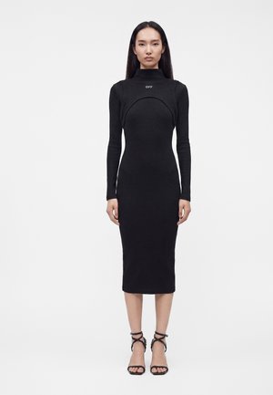 Femme debout portant une robe midi noire cintrée à manches longues, col montant et logo "OFF", associée à des sandales noires à brides.