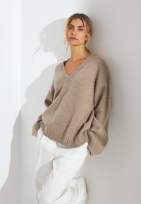 Gaiši brūns adīts džemperis ar V veida kakla izgriezumu, brīva piegriezuma un ribota apakša, apvienots ar baltām biksēm. Gluda tekstūra, ikdienišķs stils.