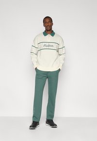 Pull à col rond crème avec accents verts, motif à carreaux et texte brodé. Porté avec un pantalon de survêtement vert et des chaussures noires.