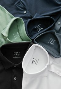 Colección de polos slim-fit en negro, blanco, verde oscuro y azul marino. Cada uno cuenta con un collar, cierre de botones y etiqueta de la marca en el interior.