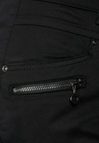 Černé kalhoty mají kapsu na zip se stříbrným kováním, ploché švy a hladkou texturu, navržené pro funkčnost a styl.