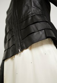 Black leather jacket with layered ruffle design, zip closure, and silver hardware; worn over a light, cream-colored garment with black dots.