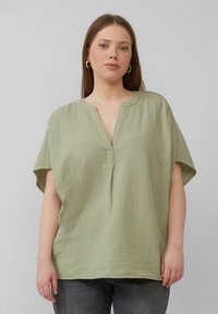 Hellgrünes, kurzärmliges Oberteil mit V-Ausschnitt, aus weichem Textil mit Struktur und lockerer Passform. Es weist ein dezentes, zerknittertes Muster auf.