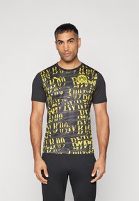 Camisa de manga corta negra y amarilla con un patrón gráfico repetido y un logo circular en la parte izquierda del pecho.