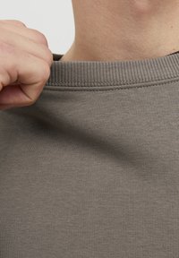 Tessuto di felpa grigia a coste con colletto a girocollo, che mostra texture e elasticità, con una mano che afferra il colletto. Messa a fuoco ravvicinata sull'area del collo.