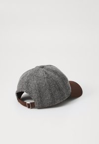 Căciulă cu model houndstooth gri, având vârful maro și curea din piele ajustabilă. Fabricată dintr-un material texturat. Vedere din spate care arată deschiderea și cureaua.