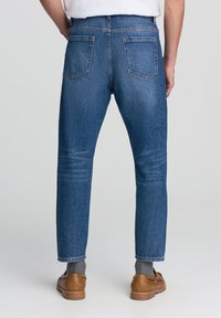 Vista trasera de una persona que lleva pantalones vaqueros azules, calcetines grises, zapatos de cuero marrón y una camisa blanca, de pie frente a un fondo liso.