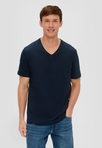 Maglietta polo blu navy con scollo a V realizzata in morbido tessuto di cotone, maniche corte, abbinata a jeans blu chiaro, in piedi davanti a uno sfondo neutro.