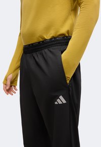 Personne portant un pantalon de course noir Adidas avec la main dans la poche et un haut jaune à manches longues avec des trous pour le pouce.