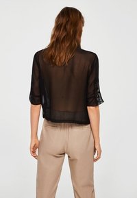 Blouse noire transparente à manches courtes, ornée d'accents en dentelle. Portée avec un pantalon beige, présentant une coupe ample et une texture transparente.