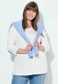 Lehký bílý top s dlouhým rukávem, výstřihem do V a vyhrnutými rukávy, doplněný modrým svetrem uvázaným přes ramena a modrými džínami.