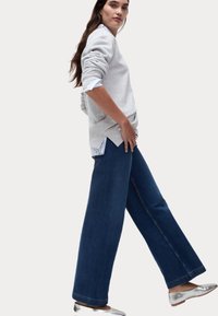Felpa grigia chiara a maniche lunghe indossata sopra una camicia azzurra con bottoni, abbinata a jeans larghi blu scuro e scarpe flat argentate.