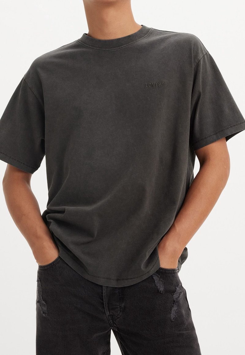 Personne portant un t-shirt Levi’s noir délavé et un jean noir déchiré, mains dans les poches avant, sur un fond uni.