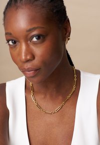 Collier en or avec des maillons allongés et une texture lisse, porté par une personne en haut blanc. Des boucles d'oreilles créoles simples complètent l'ensemble.