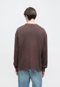 Ruda tekstūruota ilgomis rankovėmis palaidinė laisvo silueto su apvalia iškirpte, derinama su šviesiai mėlynomis džinsinėmis kelnėmis.