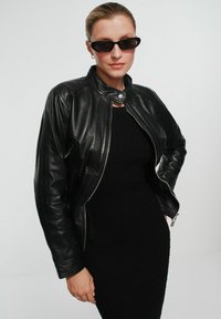 Czarna skórzana kurtka motocyklowa z wysokim kołnierzem i srebrnym zamkiem. Nosi się ją na dopasowanej czarnej sukience dzianinowej. Czarne owalne okulary przeciwsłoneczne dopełniają stylizację.