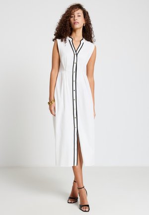 Donna che indossa un vestito midi bianco senza maniche con bordino nero e bottoni sul davanti, tacchi neri con cinturino alla caviglia, braccialetti dorati e capelli ricci.