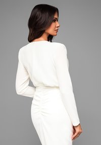 Weißes Kleid mit langen Ärmeln, das ein gerafftes Design an der Taille und einen kleinen Schlüsselloch-Verschluss auf dem Rücken aufweist. Glattes, leichtes Material.