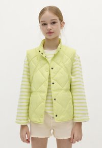Chaleco acolchado color verde claro con cierres a presión, superpuesto a una camisa de manga larga a rayas crema y lima y pantalones cortos de color beige deshilachados.