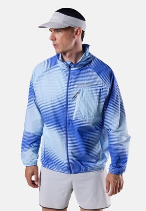 Man met een blauw-wit gestreepte jas met rits, witte korte broek en witte vizierpet, kijkt naar links tegen een effen achtergrond.