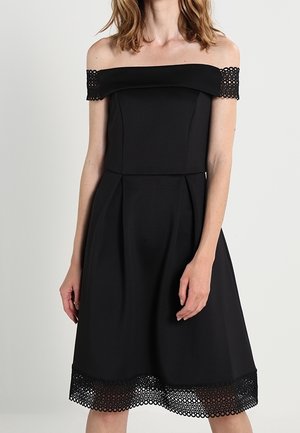 Kvinde iført en sort off-shoulder kjole med blondekant på ærmer og bund, stående mod en ensartet lys baggrund.