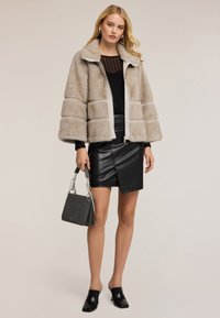 Žena v béžové umělé kožešině, černém průhledném topu, černé kožené sukni, černých podpatkových pantoflích, s šedou kabelkou, stojící na jednobarevném pozadí.