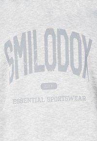 Tela gris clara con texto desvanecido "SMILODOX" en letras grandes, "2013" en un óvalo pequeño y "ESSENTIAL SPORTSWEAR" debajo.