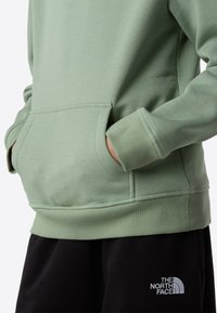 Lys grønn genser med frontlomme, ribbestrikkede mansjetter og kant, kombinert med svarte bukser med en logo på den nedre delen av benet.