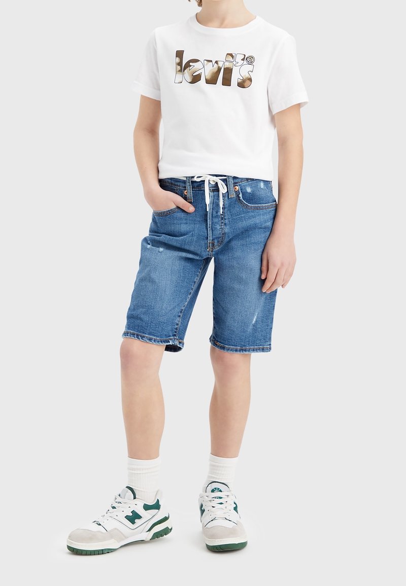 Bílý tričko s metalickým hnědým logem "Levi's", kombinované s modrými džínovými šortkami se stahovací šňůrkou a mírným opotřebením. Nosené s teniskami.