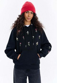 Černá mikina s kapucí zdobená květinovou výšivkou v růžové, bílé a zelené barvě. Měkký materiál, přední kapsa, nastavitelná šňůrka na kapuci, volný střih. Červená čepice.