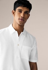 Bijela lanena košulja kratkih rukava s ovratnikom, jednim džepom na prsima i zatvaranjem na gumbe. Mekana tekstura s opuštenim krojem.