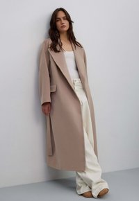 Beige lange jas met een opstaande kraag, gestructureerde schouders en zijzakken, gedragen over een wit topje en lichtgekleurde broeken.