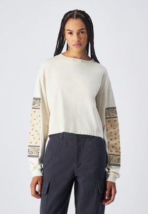 Gecombineerd crèmekleurig sweatshirt met gestreepte beige mouwen en zwarte accenten. Zachte textuur, losse pasvorm, gecombineerd met donkere cargobroeken.