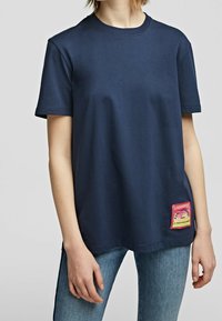 Tummansininen puuvillapaita, jossa on pyöreä pääntie, lyhyet hihat ja monivärinen kangasmerkki vasemmassa alakulmassa, jossa on tekstityksiä.