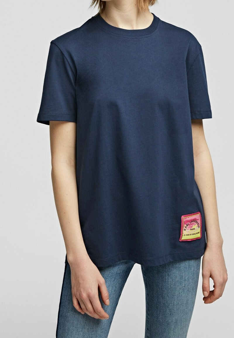 Tummansininen puuvillapaita, jossa on pyöreä pääntie, lyhyet hihat ja monivärinen kangasmerkki vasemmassa alakulmassa, jossa on tekstityksiä.