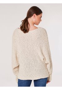 Kremowy, oversize sweter o teksturowanym splocie, z opuszczonymi ramionami i ryflowanymi mankietami. Noszony na dżinsach, prezentujący luźny krój.