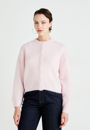 Cardigan flou rose clair avec un col rond et un système de fermeture à boutons. Il présente des manches longues et un ourlet court, associé à un denim foncé.