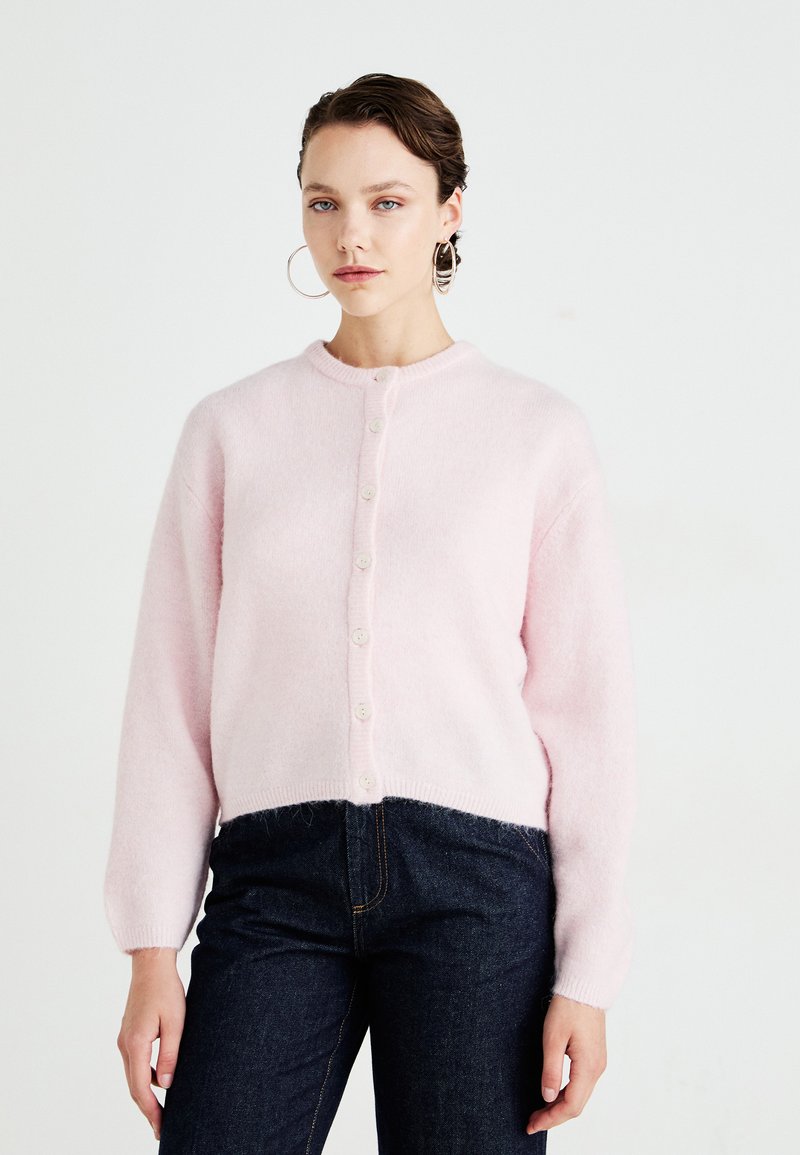 Απαλή ροζ, φουντωτή κάρντιγκαν με στρογγυλή λαιμόκοψη και κλείσιμο με κουμπί. Διαθέτει μακριά μανίκια και κοντό τελείωμα, συνδυασμένο με σκούρο τζιν.