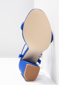 Sininen mokkanahkainen sandaali korkealla korolla, jossa on sileä ruskea kumipohja ja ohuet hihnat. Suunnittelu on avointoimitteinen ja virtaviivainen.