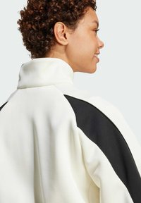 White jacket with a high collar, featuring black accents on the shoulders, made from a smooth fabric with a relaxed fit and soft texture.